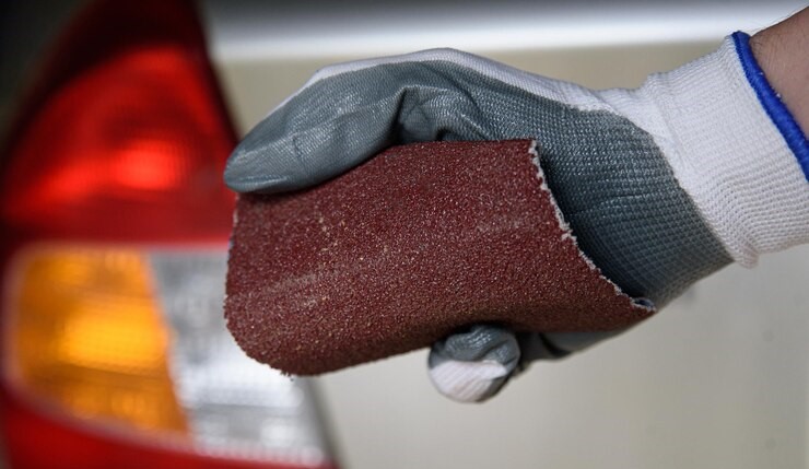 A Glove Holding a Piece of Sandpaper in Front of a white Car that is out of Focus